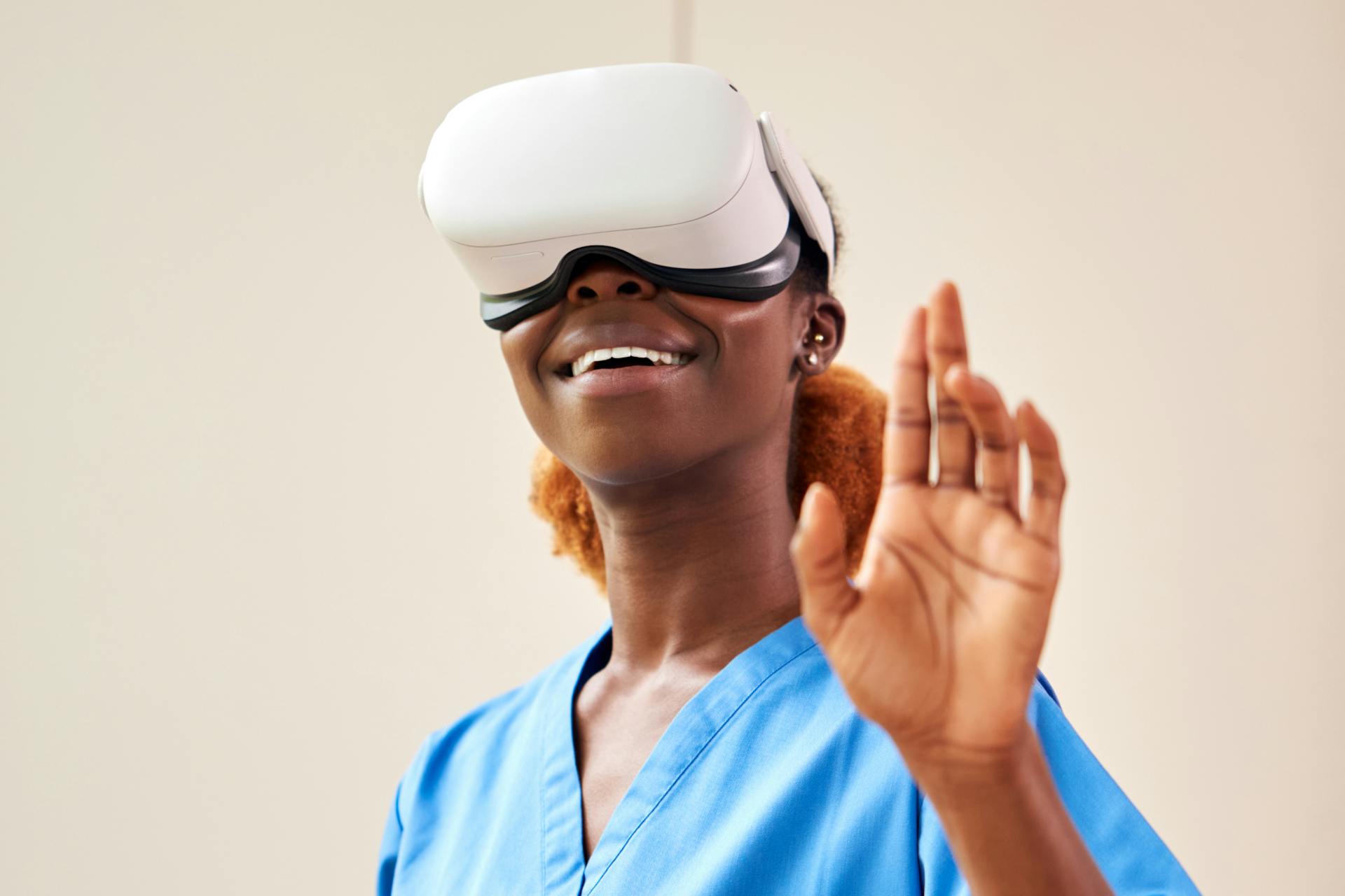A headshot of woman who is wearing a VR headset, she's smiling and raising one hand up.